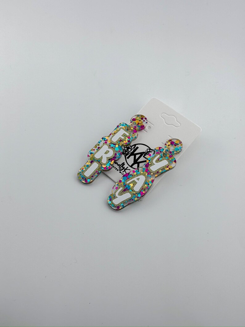 K&ouml;nnte beinhalten: Ein Paar H&auml;ngeohrringe mit dem Wort "FRIDAY" in wei&szlig;en Buchstaben auf einem goldenen, blauen, rosa und gr&uuml;nen Glitzerhintergrund.