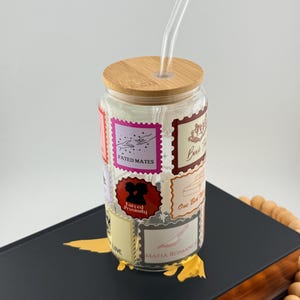 Puede incluir: Un vaso de vidrio transparente con tapa de bambú y pajita de vidrio. El vaso está decorado con varios gráficos estilo sello postal, incluyendo los textos "Fated Mates" y "Mafia Romance". El vaso está sobre un libro negro.