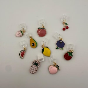 May include: A selection of fruit-shaped charms, such as a strawberry, avocado, peach, lemon, blueberry, watermelon, pineapple, and cherry. Each charm is brightly coloured with gold-toned details and attached to a clear clip.