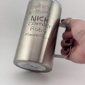 May include: A silver stainless steel travel mug with a handle. The mug has the names "Sydney", "Anthony", "Nick", "Camden", "Ruby", and "Mackenzie" written on it.