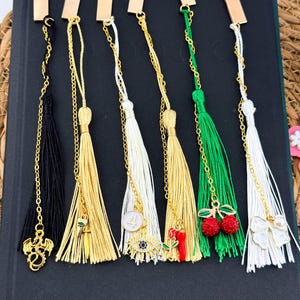 May include: A collection of decorative bookmarks with gold-colored metal tops and tassels in various colors. Each bookmark features a unique charm, including a dragon, a dagger, a number 4, an eye, a rose, cherries, and a bow.