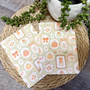 May include: Two rectangular cards with a light green background and a repeating pattern of pumpkins, bows, and floral elements. The cards are placed on a woven, circular placemat, with a small white pot of greenery in the background.
