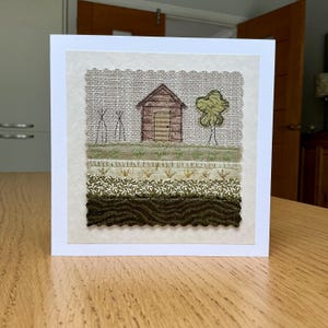 May include: A square greeting card featuring a fabric collage of a landscape. The scene includes a brown shed, a green tree, and two thin black structures on a beige linen background. Below, layers of green and brown fabric depict textured ground.