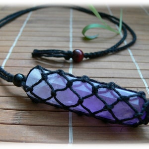 May include: A purple crystal encased in a black macrame net, hanging from a black cord with a wooden bead. The crystal is a long, thin shape and is suspended from the cord by a black bead.