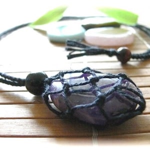 May include: A purple amethyst stone is encased in a black macrame net, hanging from a black cord necklace with a black bead. The necklace is on a bamboo mat.