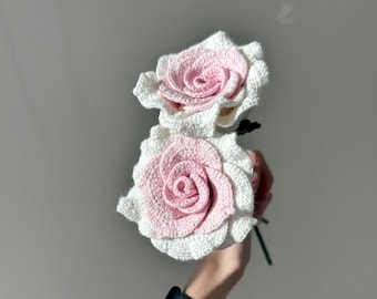 May include: Two crocheted roses, one white and one pink, are held in a hand. The roses are made with a delicate, layered design.