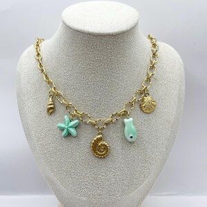 Puede incluir: Un collar de cadena dorada con una variedad de dijes con temática marina, incluyendo una estrella de mar, una concha y un pez. Los dijes están pintados en un color verde claro.