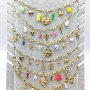 Peut inclure: Une collection de cinq colliers à chaînes dorées, chacun orné d'un assortiment de breloques colorées incluant des soleils, étoiles, cœurs, coquillages, perles et diverses figurines miniatures, présentés sur un présentoir en velours gris clair.