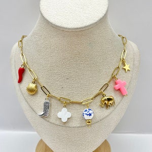 Puede incluir: Un collar de cadena dorada con múltiples dijes, que incluyen un chile rojo, una concha, una bota vaquera plateada, un trébol blanco, una cuenta con estampado azul y blanco, una cruz rosa, un elefante dorado y una estrella dorada.