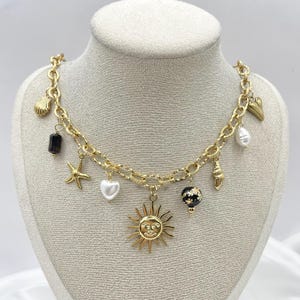 Puede incluir: Collar de cadena dorada con múltiples dijes, incluyendo un sol, un corazón, una concha, una estrella de mar y una cuenta blanca y negra.