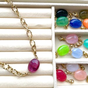 Peut inclure: Un collier à chaîne dorée avec une pierre ovale magenta, accompagné d'une collection de breloques en pierre ovale de différentes couleurs, chacune équipée d'un fermoir mousqueton doré pour permettre de les attacher facilement.