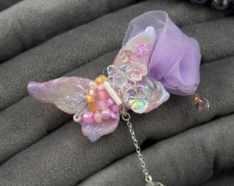 May include: A handcrafted butterfly brooch with iridescent wings, adorned with pink and purple beads, and a dangling crystal charm. The brooch features a delicate chain and a sheer lavender fabric accent. A dark gray beaded necklace is in the background.