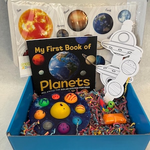 Puede incluir: Una caja azul llena de artículos coloridos para una actividad de aprendizaje sobre el espacio. La caja contiene un libro titulado "My First Book of Planets", un juguete pop-it con forma de sistema solar, una hoja de pegatinas con planetas y una página para colorear de un OVNI.