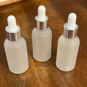 May include: Three frosted glass dropper bottles with white droppers and silver collars. The bottles are arranged on a wooden surface. The bottles are empty and ready to be filled with product.