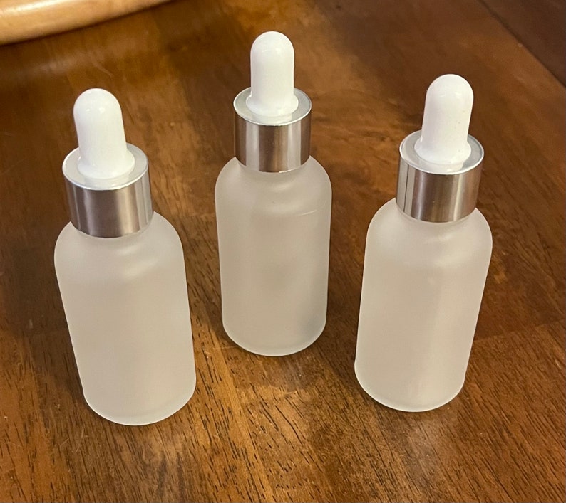 May include: Three frosted glass dropper bottles with white droppers and silver collars. The bottles are arranged on a wooden surface. The bottles are empty and ready to be filled with product.