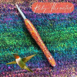 May include: A sparkly, orange and red crochet hook with a silver metal tip, resting on a colorful knitted fabric. The text "Ruby-Throated" is in a red oval. A hummingbird is in the lower left corner.