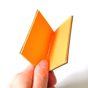 May include: A small, open book with bright orange pages and black edges is held in a hand against a white background. The book is open, displaying the interior pages. The cover and pages are a vibrant orange.