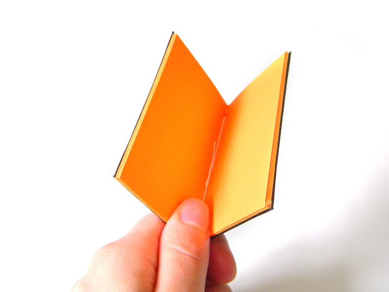 May include: A small, open book with bright orange pages and black edges is held in a hand against a white background. The book is open, displaying the interior pages. The cover and pages are a vibrant orange.