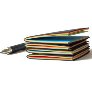 May include: A stack of colorful sticky note pads with black edges, featuring a rainbow of colors. A silver and black pen rests beside the pads, suggesting a writing or note-taking theme. The pads are on a white background.