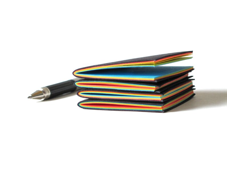 May include: A stack of colorful sticky note pads with black edges, featuring a rainbow of colors. A silver and black pen rests beside the pads, suggesting a writing or note-taking theme. The pads are on a white background.