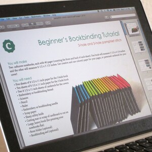 May include: A laptop displays a bookbinding tutorial titled "Beginner's Bookbinding Tutorial." The tutorial lists materials needed, including paper, thread, and scissors. A stack of colorful paper is shown. The laptop is open on a desk.