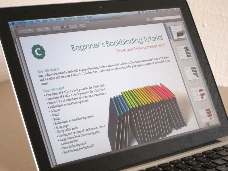 May include: A laptop displays a bookbinding tutorial titled "Beginner's Bookbinding Tutorial." The tutorial lists materials needed, including paper, thread, and scissors. A stack of colorful paper is shown. The laptop is open on a desk.