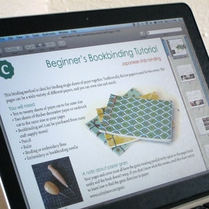 May include: A laptop screen displays a bookbinding tutorial titled "Beginner's Bookbinding Tutorial" with "Japanese stab binding" as the method. The tutorial includes a list of materials and instructions, along with images of finished bookbinding projects. The screen also shows a preview of the tutorial.