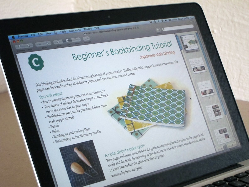 May include: A laptop screen displays a bookbinding tutorial titled "Beginner's Bookbinding Tutorial" with "Japanese stab binding" as the method. The tutorial includes a list of materials and instructions, along with images of finished bookbinding projects. The screen also shows a preview of the tutorial.
