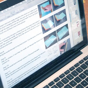 May include: A laptop displaying instructions for a craft project. The screen shows step-by-step images and text, including instructions for sewing a book. The laptop is open on a wooden surface, with the keyboard visible.
