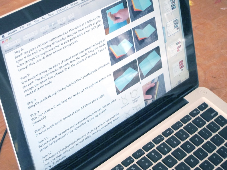 May include: A laptop displaying instructions for a craft project. The screen shows step-by-step images and text, including instructions for sewing a book. The laptop is open on a wooden surface, with the keyboard visible.