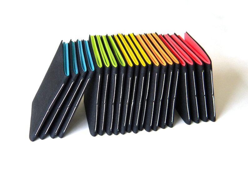 May include: A series of black, accordion-style notebooks with colorful edges. The edges transition from blue to green, yellow, orange, and red. Each notebook is held together with white stitching.