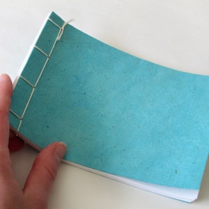 May include: A small, handmade notebook with a turquoise cover and white interior pages. The binding is visible, with cream-colored thread stitching the pages together. The notebook is held open, showcasing the textured paper.