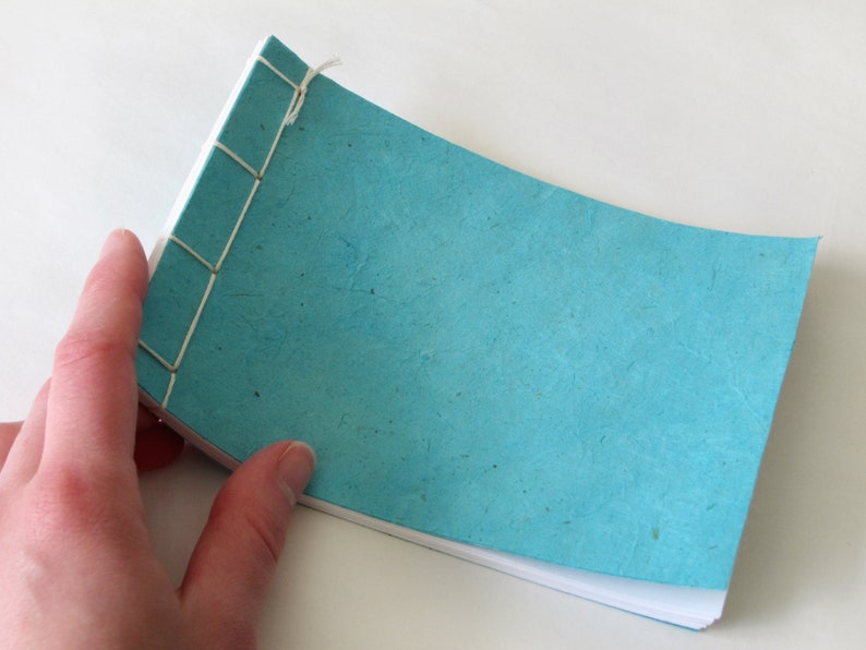 May include: A small, handmade notebook with a turquoise cover and white interior pages. The binding is visible, with cream-colored thread stitching the pages together. The notebook is held open, showcasing the textured paper.