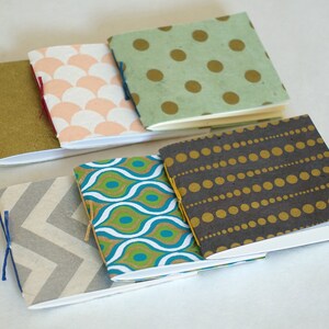 May include: A collection of small, handmade notebooks with patterned covers. The covers feature designs like gold polka dots on green, a chevron pattern, and a teal and white design. Each notebook is bound with colorful thread.