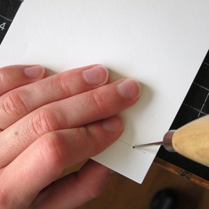 May include: A close-up of a hand using a wooden handled awl to pierce a white piece of paper. The paper is on a black cutting mat with a ruler. The awl is silver and the paper is white.