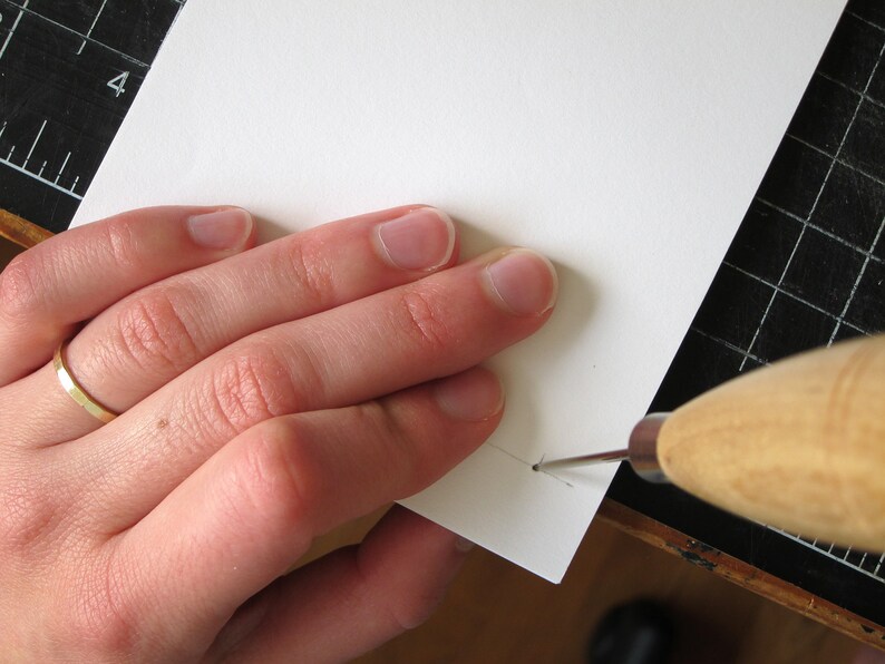 May include: A close-up of a hand using a wooden handled awl to pierce a white piece of paper. The paper is on a black cutting mat with a ruler. The awl is silver and the paper is white.