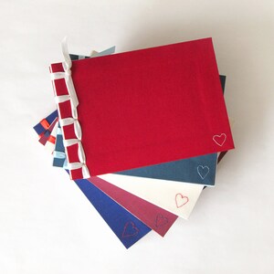 May include: A stack of handmade journals with red, blue, and white covers. Each journal has a heart embroidered in the corner. The spines are bound with a decorative ribbon. The journals are a mix of sizes and colors.