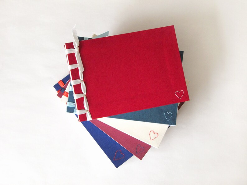 May include: A stack of handmade journals with red, blue, and white covers. Each journal has a heart embroidered in the corner. The spines are bound with a decorative ribbon. The journals are a mix of sizes and colors.