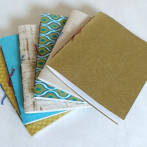 May include: A collection of handmade notebooks with various patterned covers. The notebooks are bound with colorful thread and feature covers in shades of gold, teal, blue, and patterned fabric. The notebooks are stacked in a fan shape.