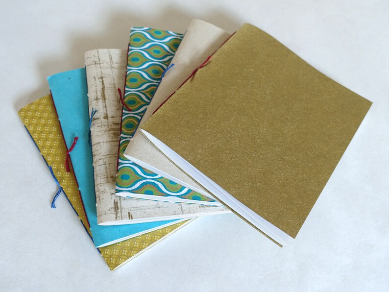 May include: A collection of handmade notebooks with various patterned covers. The notebooks are bound with colorful thread and feature covers in shades of gold, teal, blue, and patterned fabric. The notebooks are stacked in a fan shape.