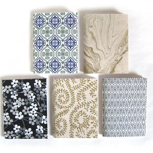 May include: Five small notebooks with patterned covers. Designs include a blue and green geometric pattern, a beige marbled design, a black floral print, a gold leaf pattern, and a blue and white scroll design. The notebooks are rectangular and arranged on a white surface.