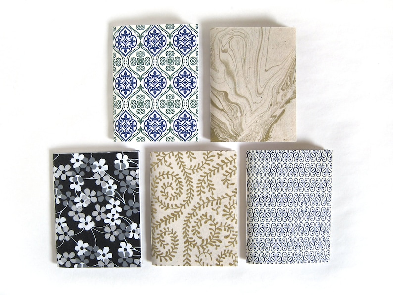 May include: Five small notebooks with patterned covers. Designs include a blue and green geometric pattern, a beige marbled design, a black floral print, a gold leaf pattern, and a blue and white scroll design. The notebooks are rectangular and arranged on a white surface.
