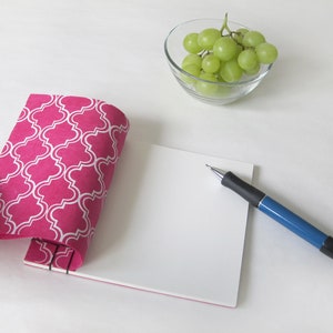 May include: A pink notebook with a white quatrefoil pattern is held open, revealing blank white pages. A blue and black pen rests on the pages. A small glass bowl of green grapes sits nearby. The notebook is bound with black staples.