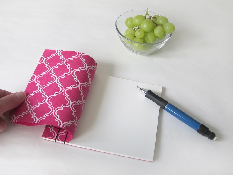 May include: A pink notebook with a white quatrefoil pattern is held open, revealing blank white pages. A blue and black pen rests on the pages. A small glass bowl of green grapes sits nearby. The notebook is bound with black staples.