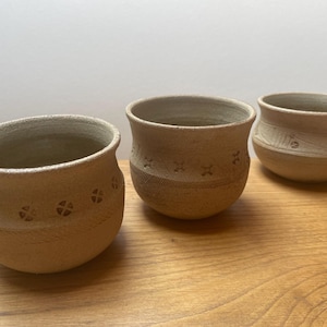 Może przedstawiać: Trzy ręcznie robione ceramiczne miski w naturalnym beżowym kolorze. Każda miska ma unikalny wzór, w tym wzory w kształcie krzyża i teksturowane paski. Miski są prezentowane na drewnianej powierzchni, prezentując ich rustykalny urok i kunszt.
