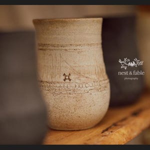 May include: A beige ceramic tumbler with a textured, speckled surface featuring horizontal incised lines and a small cross-shaped stamp. The vessel has a slightly tapered cylindrical shape and includes the text nest & fable photography.