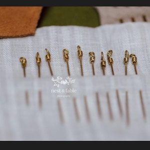 May include: A collection of gold-toned metal needles with looped eyes arranged in a row on a white textured fabric. The needles are positioned vertically, with their pointed ends extending downward into the fabric. The text nest & fable is visible.