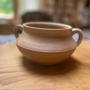 May include: A light brown ceramic pot with two curved handles. The pot has a rounded body with horizontal ridges and a wide opening. The pot is on a wooden surface.