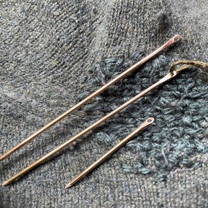 Puede incluir: Tres agujas de coser metálicas de diferentes tamaños descansan sobre una tela gris texturizada. Las agujas son de un color cobre cálido, con pequeños ojales en la parte superior. Un hilo verde está unido a una aguja.