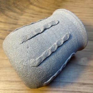 Op de afbeelding: Een grijze keramische beker met een textuuroppervlak en twee verhoogde, golvende banden. De handgemaakte beker heeft een licht uitlopende rand en staat op een houten achtergrond. De beker is ongeveer 10 cm hoog.